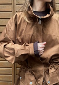 Brun semsket jakke med høy krage, innsvingt midje, store frontlommer og mørke mansjetter. Har glidelås og trykknapper.