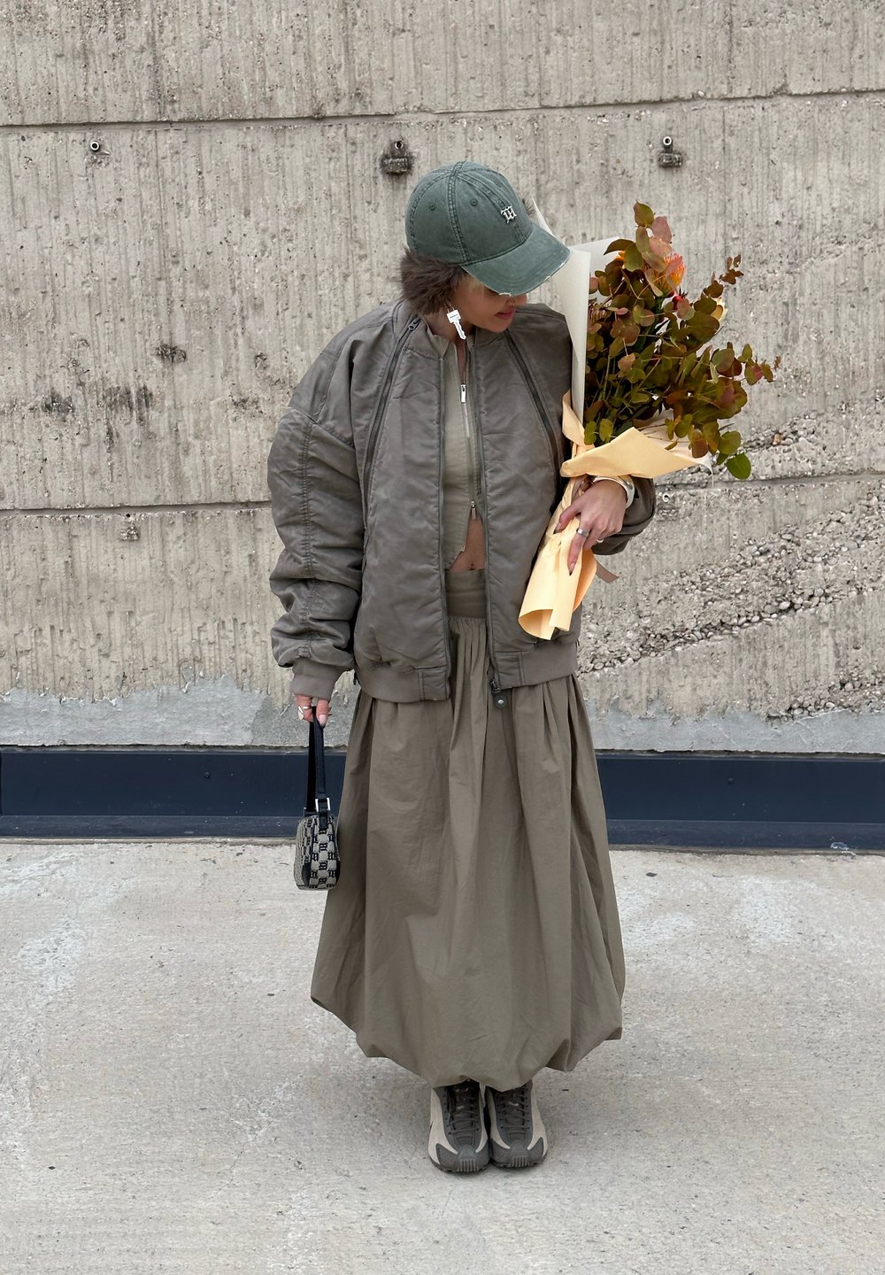 Olive green bomber jacket, cropped beige top, flowing long skirt, grey trainers, holding a floral bouquet wrapped in pale yellow. Small handbag.
