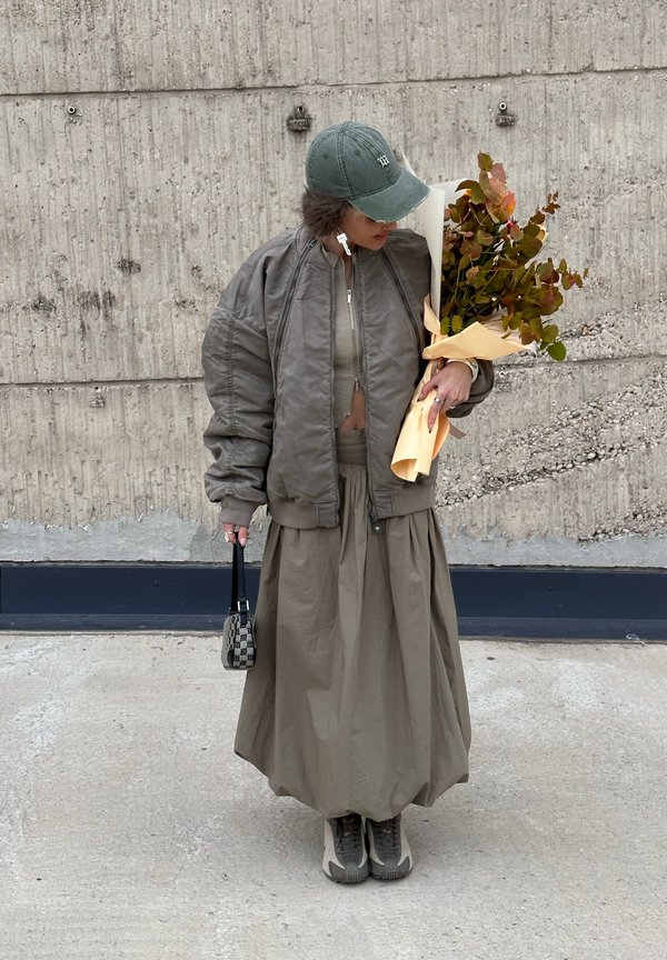 Olive green bomber jacket, cropped beige top, flowing long skirt, grey sneakers, holding a floral bouquet wrapped in light yellow. Small handbag.
