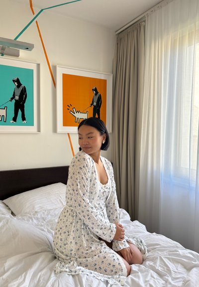 Vestido floral blanco con mangas largas, tela texturizada. Cama con sábanas blancas, dos obras de arte enmarcadas en la pared, cortinas beige y luz solar brillante.