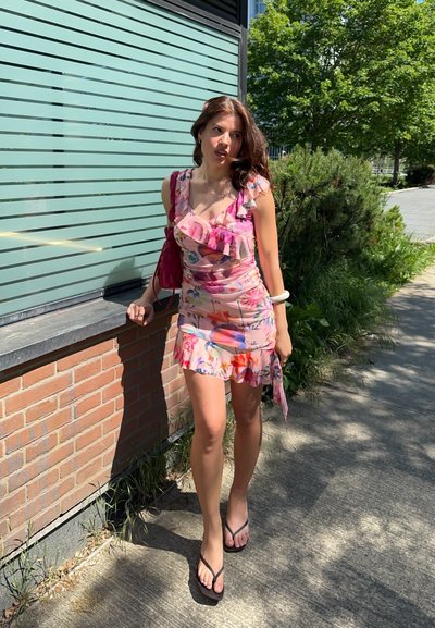 Mujer caminando por la acera con un vestido rosa con flores, sosteniendo un bolso a juego, cerca de una pared de ladrillos y plantas verdes a la luz del día.