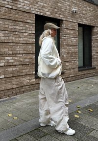 Balta bumbiera jaka, vaļīgas bēša krāsas bikses un balti sporta apavi. Zaļgani olīvu cepure. Teksturētas ķieģeļu sienas fons ar lapotni uz ietves.