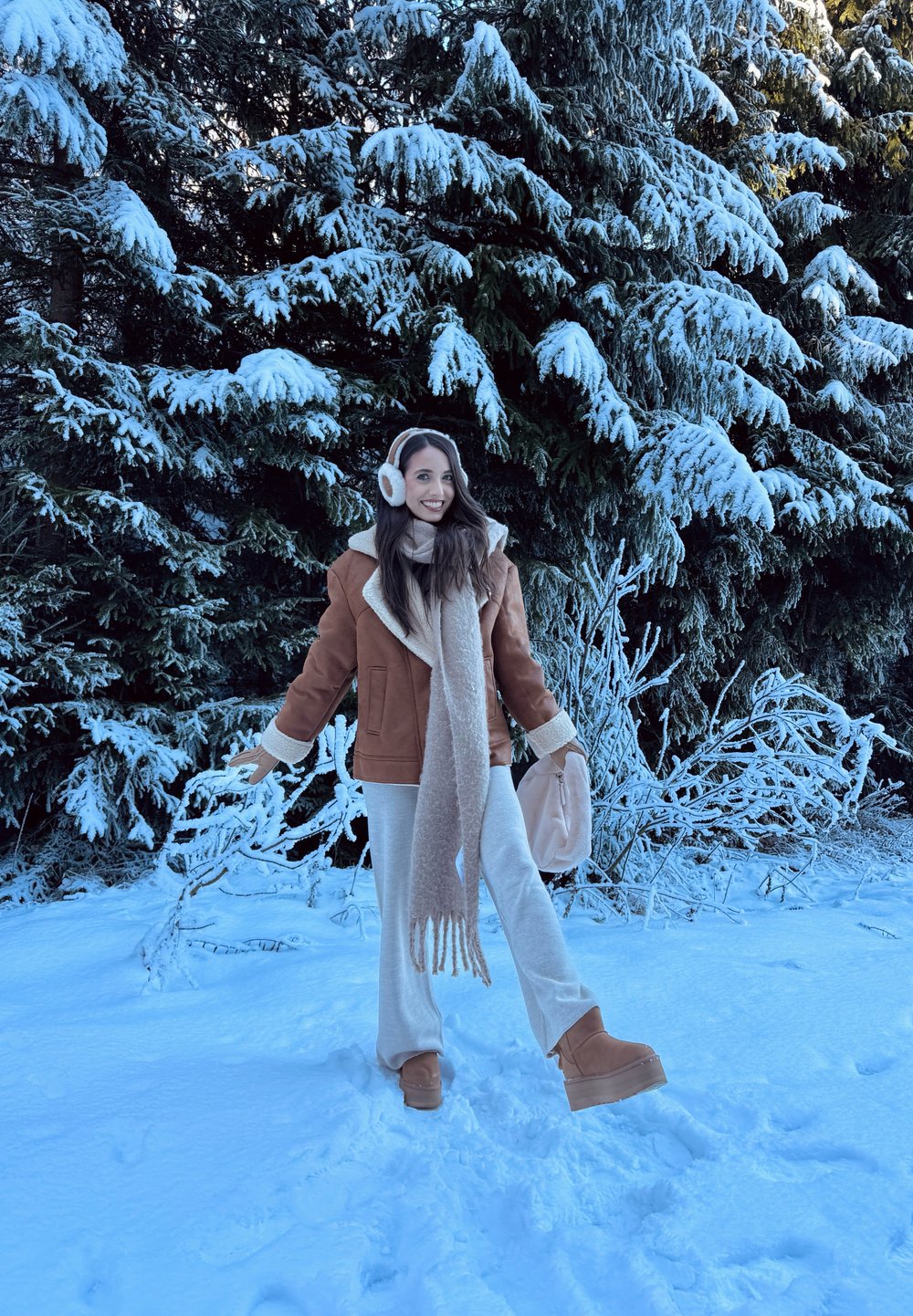 Jachetă din blană maro, eșarfă crem, pantaloni bej și cizme de iarnă maronii. Copaci verzi acoperiți de zăpadă în fundal. Pământ acoperit de zăpadă strălucitor.