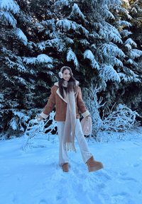 Jachetă din blană maro, eșarfă crem, pantaloni bej și cizme de iarnă maronii. Copaci verzi acoperiți de zăpadă în fundal. Pământ acoperit de zăpadă strălucitor.