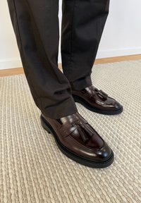 Mocassins à pampilles en cuir marron foncé portés avec un pantalon noir, posés sur un tapis beige texturé.