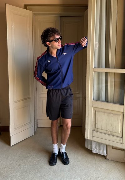 Chaqueta deportiva azul marino con franjas rojas, pantalones cortos negros, calcetines blancos y mocasines negros. De pie frente a un marco de puerta.