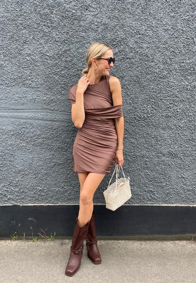 Vestido mini marrón con fruncidos y un diseño de manga asimétrica; combinado con botas marrones hasta la rodilla y un bolso de mimbre. Fondo de pared texturizada gris.