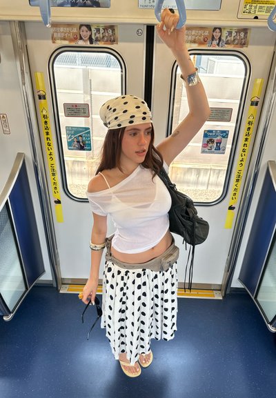 Una persona dentro de un tren, vistiendo un top blanco de hombros descubiertos, un pañuelo de lunares en la cabeza y una falda plisada a rayas en blanco y negro con lunares, sosteniendo un bolso.