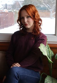 Jeune femme aux cheveux roux bouclés, portant un pull violet foncé et un jean bleu, assise à l'intérieur près d'une fenêtre avec de la neige à l'extérieur et une plante verte à proximité.