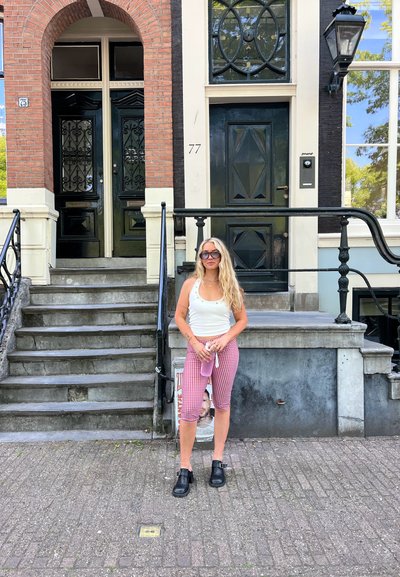 Una mujer se encuentra en unas escaleras de piedra al lado de una casa de ladrillos rojos y puertas negras. Lleva una camiseta de tirantes blanca y leggings a cuadros rosas.