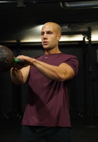 Άνδρας με ξυρισμένο κεφάλι που σηκώνει μια καμπάνα στο γυμναστήριο με χαμηλό φωτισμό, φοράει μπλούζα με κοντά μανίκια μπορντό και είναι συγκεντρωμένος στην άσκηση.