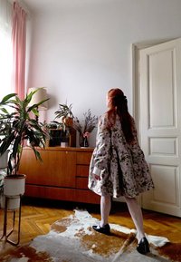 Mētelis ar ziedu rakstu, plandošām piedurknēm, baltām aprocēm un teksturētu virsmu. Telpā ir koka kumode, augi un brūns govs ādas paklājs.