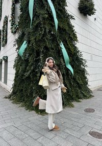 Femme en manteau beige, écharpe, cache-oreilles et moufles tenant des sacs de courses, posant avec une jambe levée près d'un grand sapin de Noël décoré en plein air.