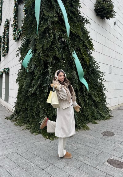 Mujer con abrigo beige, bufanda, orejeras y mitones sosteniendo bolsas de compras, posando con una pierna levantada cerca de un gran árbol de Navidad decorado al aire libre.