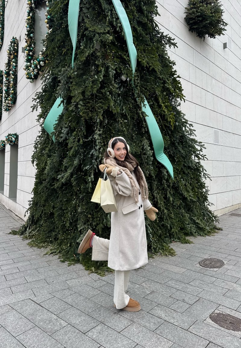 Femme en manteau beige, écharpe, cache-oreilles et moufles tenant des sacs de courses, posant avec une jambe levée près d'un grand sapin de Noël décoré en plein air.