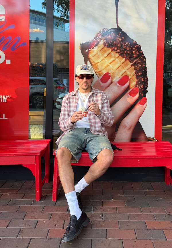 Uomo con occhiali da sole e cappellino che mangia un gelato seduto su una panchina rossa di fronte a un grande cartellone pubblicitario di un cono gelato.