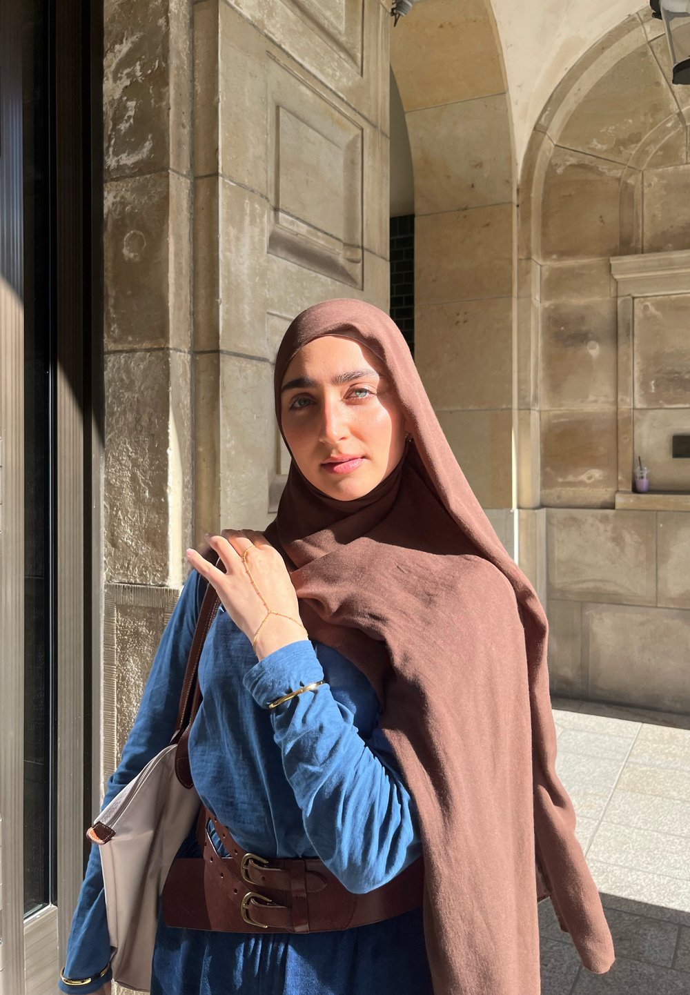 Un hijab marrone si libra su una camicia blu a maniche lunghe, con una cintura marrone. L'ambientazione include architettura in pietra con una luce solare morbida.