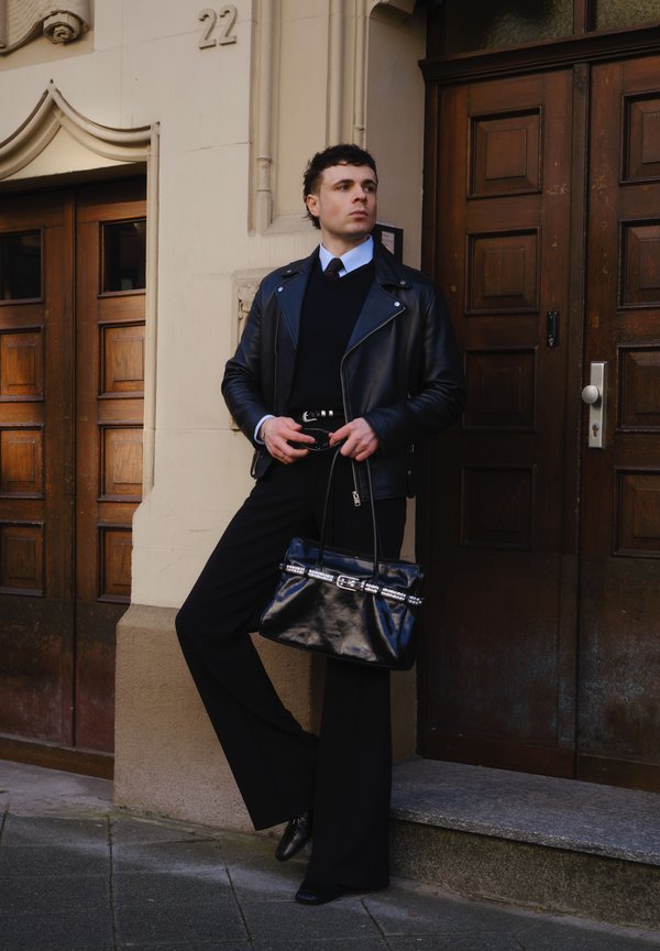 Jonge man in een zwarte leren jas en wijde broek leunt tegen een beige gebouw met houten deuren, terwijl hij een zwarte handtas en een zonnebril vasthoudt.