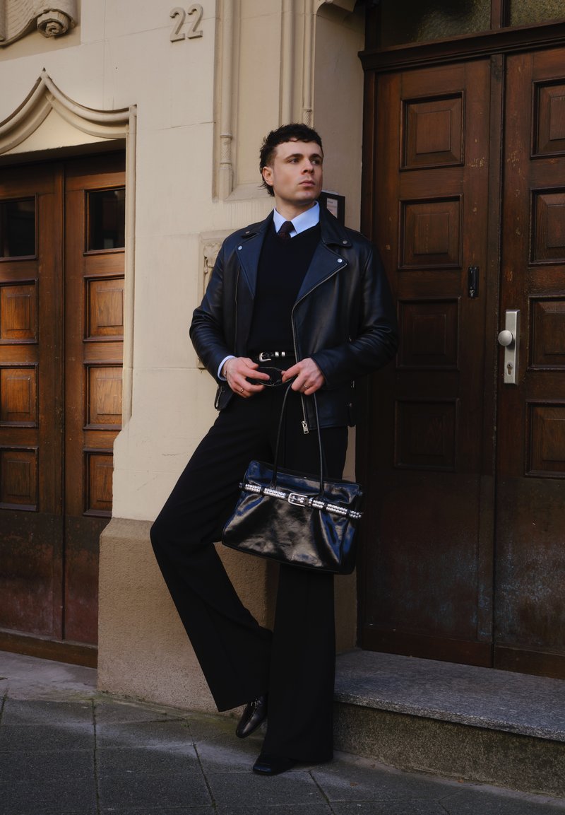 Mladić u crnoj kožnoj jakni i hlačama širokih nogavica naslonjen je na bež zgradu s drvenim vratima, držeći crnu torbu i sunčane naočale.