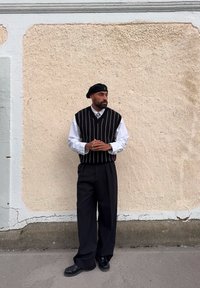 Svart- og hvitstripete vest over en hvit skjorte, sammen med høyt liv svarte bukser og svarte sko, mot en teksturert beige vegg.
