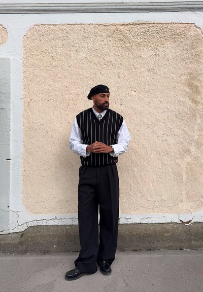 Chaleco de rayas blancas y negras sobre una camisa blanca, combinado con pantalones negros de cintura alta y zapatos negros, frente a una pared beige texturizada.