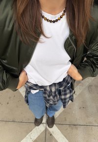 Veste bomber verte, t-shirt blanc, chemise en flanelle nouée à la taille, jean bleu, bottines marron et collier en perles.