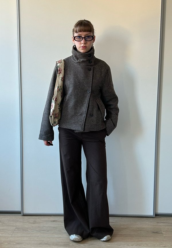 Jeune personne portant une veste grise boutonnée, un pantalon large noir à fines rayures, des baskets blanches, des lunettes noires, portant un sac fourre-tout fleuri à l'épaule gauche.