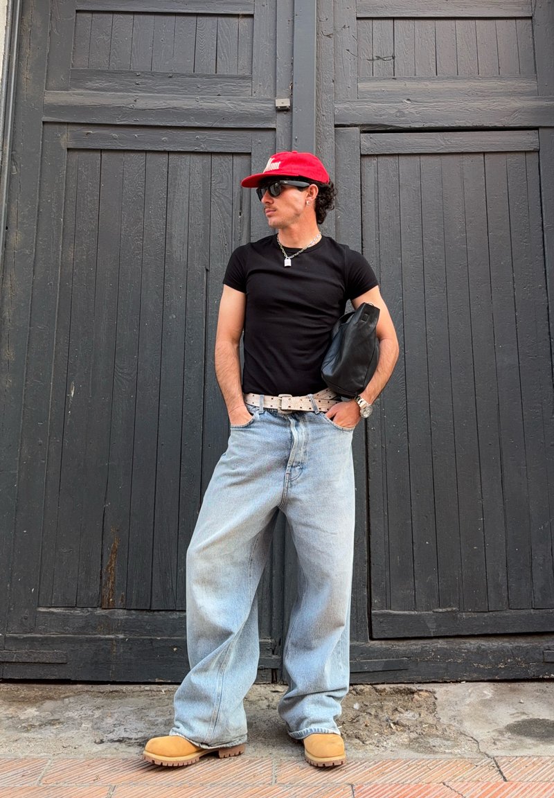 Homme portant une casquette rouge, un t-shirt noir, un jean bleu clair oversized, des bottes couleur cuir clair et des lunettes de soleil, debout les mains dans les poches devant des portes en bois sombre.