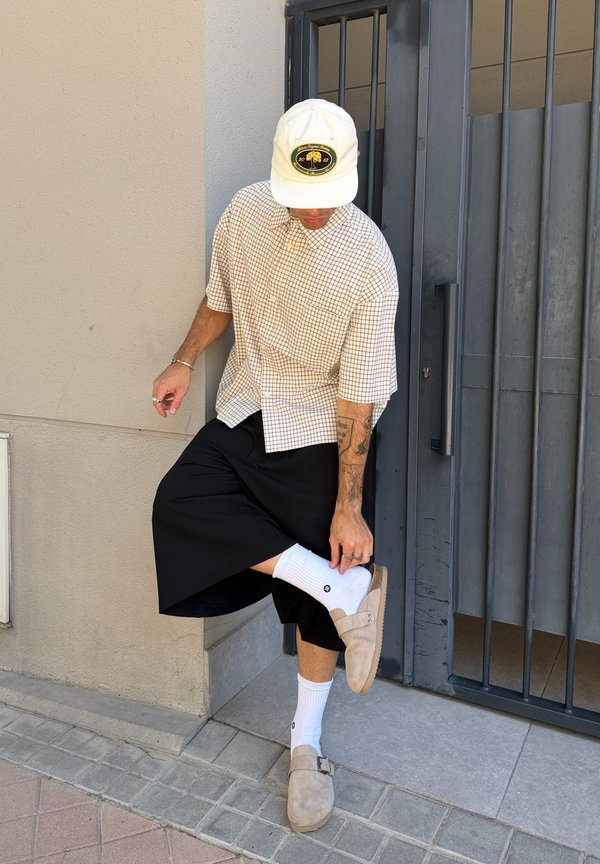 Person trägt eine beige Kappe, kariertes Hemd, schwarze weite Hosen, weiße Socken und beige Slipper und richtet ihren Schuh nahe einem Metalltor.