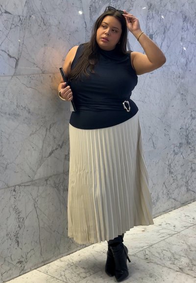 Mujer con top negro sin mangas y falda plisada beige ajustándose las gafas de sol en una habitación con paredes de mármol, sosteniendo un bolso de mano negro y usando botas negras.