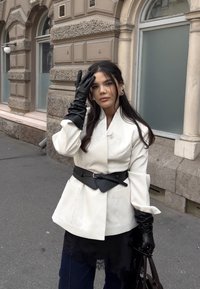 Cremefarbener, maßgeschneiderter Blazer mit einem taillierten Gürtel, kombiniert mit schwarzen Handschuhen und einem mehrlagigen Rock mit Spitzen-Details. Dunkle Denim-Hose und Tasche.