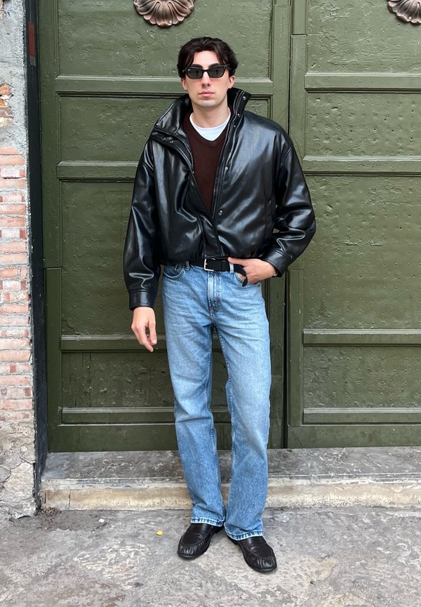Joven con chaqueta de cuero negra, gafas de sol oscuras, vaqueros azules y zapatos negros, de pie frente a una gran puerta verde.