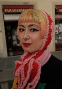 Persona con capelli corti color pesca e giallo pastello, che indossa un foulard lavorato a maglia rosso e rosa e un rossetto rosso intenso, in piedi all'aperto.