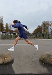 Persona con giacca con cappuccio blu navy e pantaloncini salta oltre una barriera di cemento con accenti arancioni, indossando scarpe sportive bianche e calzini.