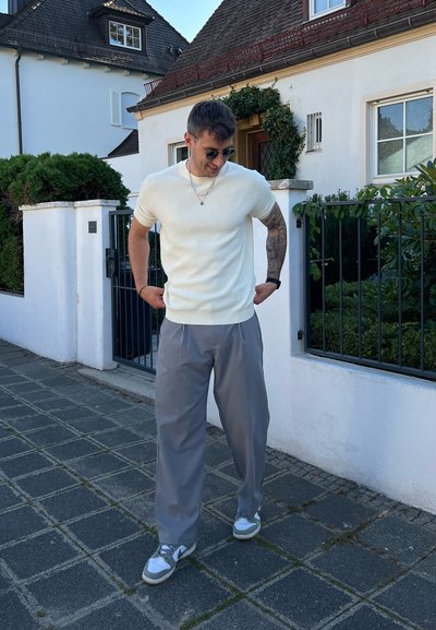 Suéter de manga corta de rayas en color crema claro, pantalones sueltos grises y zapatillas grises con acentos claros. De pie sobre un camino de piedras cerca de una casa.