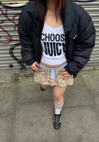 Casaco puffer preto, top branco com texto "ESCOLHE SUCO", saia mini xadrez com detalhe de botão azul, sapatos pretos e meias listradas.