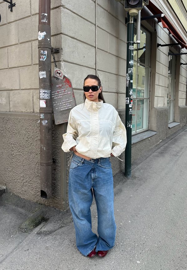 Kvinne med solbriller, hvit jakke og vide blå jeans står med hendene i lommene på en urbane fortau nær en steinbygning.