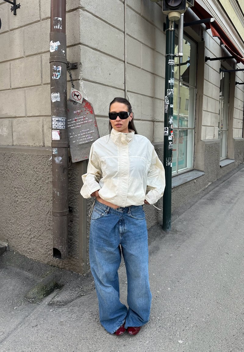 Woman wearing sunglasses, white jacket, and wide blue jeans stands with hands in pockets on urban sidewalk near stone building.