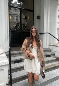 Brun, oversized jakke over en hvid kjole med blondekant; iført mørke solbriller. Står på stenskridt med en detaljeret sort dør bagved.