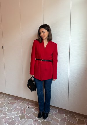 Blazer rojo a medida con mangas largas y escote en V, ceñido con un cinturón negro. Combinado con vaqueros oscuros y un bolso negro. Fondo texturizado.