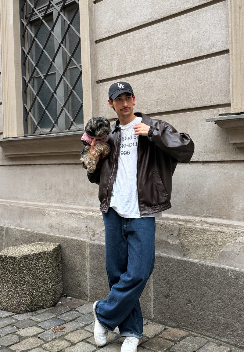 Braune Leder-Bomberjacke, weißes T-Shirt mit Aufschrift, blaue Jeans mit weitem Bein, weiße Turnschuhe, hält einen kleinen Hund in einem rosa Geschirr.