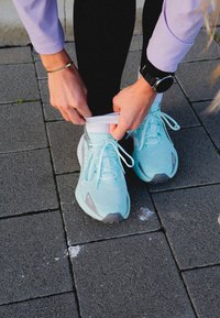 Light blue trainers with a textured fabric upper, grey accents, and laces; worn with a white sock and black leggings on a stone surface.