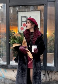 Long black coat with a faux fur collar, maroon beret, burgundy gloves, and a skirt. Holding a potted plant and a coffee cup. Urban background.