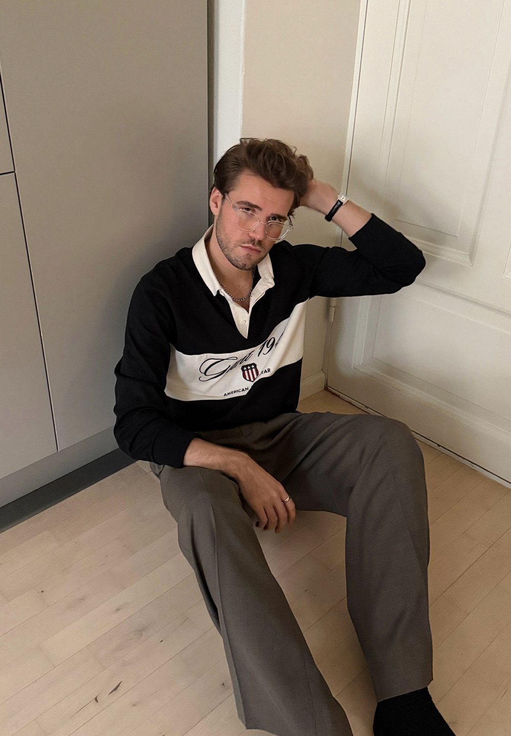 Polo à manches longues noir et blanc avec des rayures horizontales et un logo, associé à un pantalon marron clair, assis sur un sol en bois.