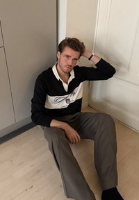 Polo à manches longues noir et blanc avec des rayures horizontales et un logo, associé à un pantalon marron clair, assis sur un sol en bois.