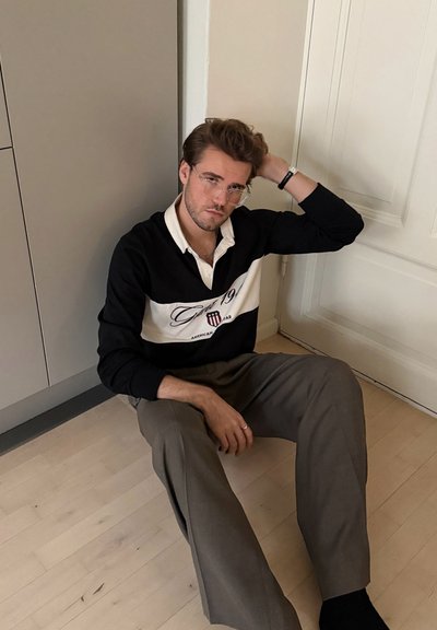 Polo de manga larga en blanco y negro con rayas horizontales y logotipo, combinado con pantalones marrón claro, sentado en un suelo de madera.