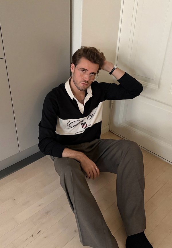 Zwart-wit polo met lange mouwen met horizontale strepen en logo, gecombineerd met lichtbruine broek, zittend op een houten vloer.