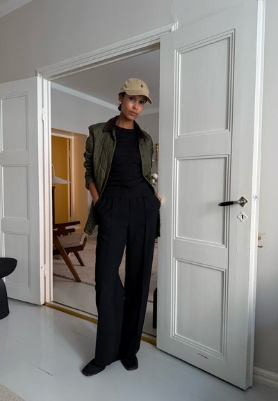 Chaqueta acolchada verde oliva con cuello marrón, suéter negro y pantalones anchos negros. Gorra beige y zapatos negros, de pie en una puerta.