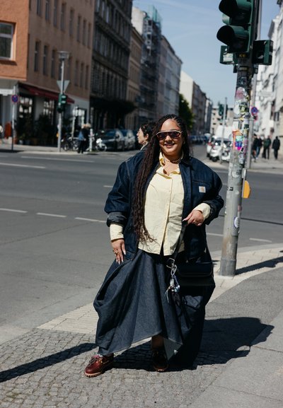 Mujer con trenzas largas que lleva gafas de sol, chaqueta vaquera, camiseta amarilla y falda larga, de pie en una esquina de una calle de la ciudad cerca de un semáforo.