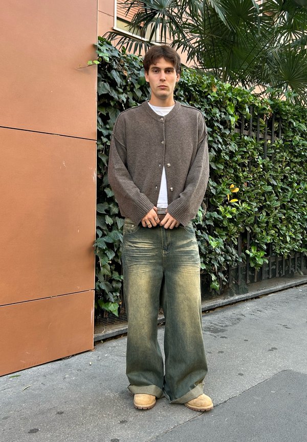Gray knitted cardigan with buttons, white t-shirt underneath, wide faded blue jeans, and beige boots; standing against greenery and a brown wall.